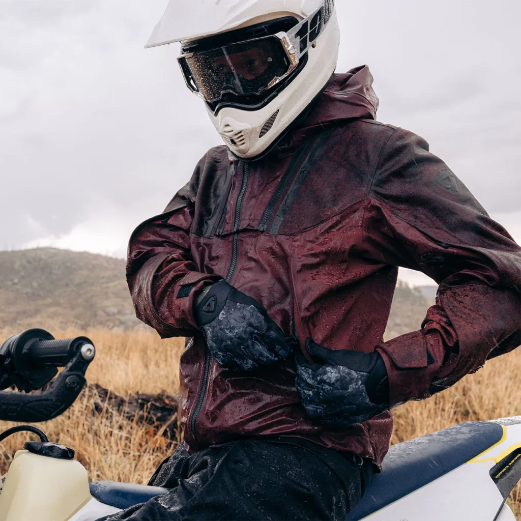 A rider on a dual-sport motorcycle navigating a muddy trail while wearing the Rev'It Component 2 H2O jacket.